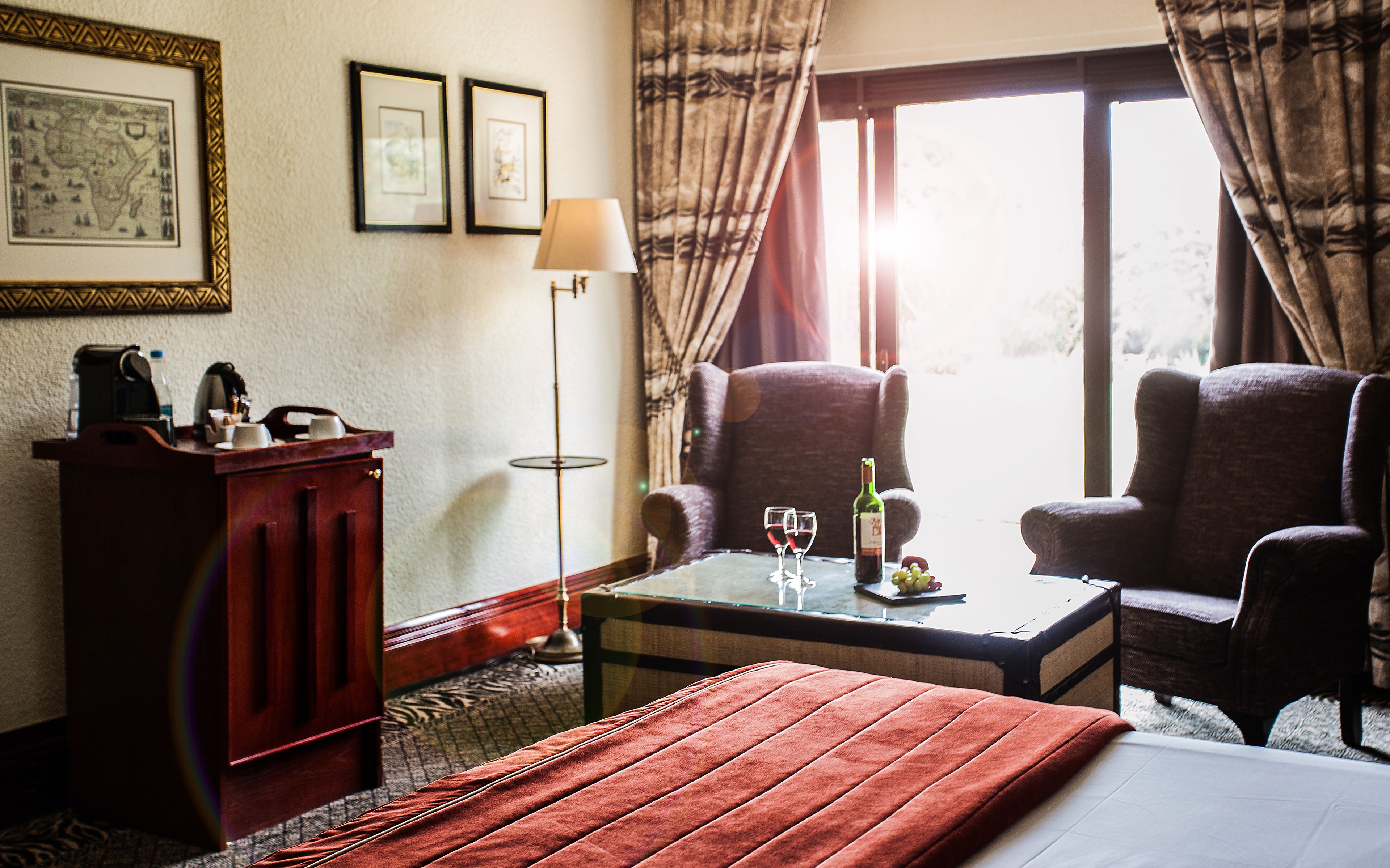 A hotel room with a bed, two armchairs, a coffee table with a bottle and glasses, a desk with a lamp, and a map on the wall.