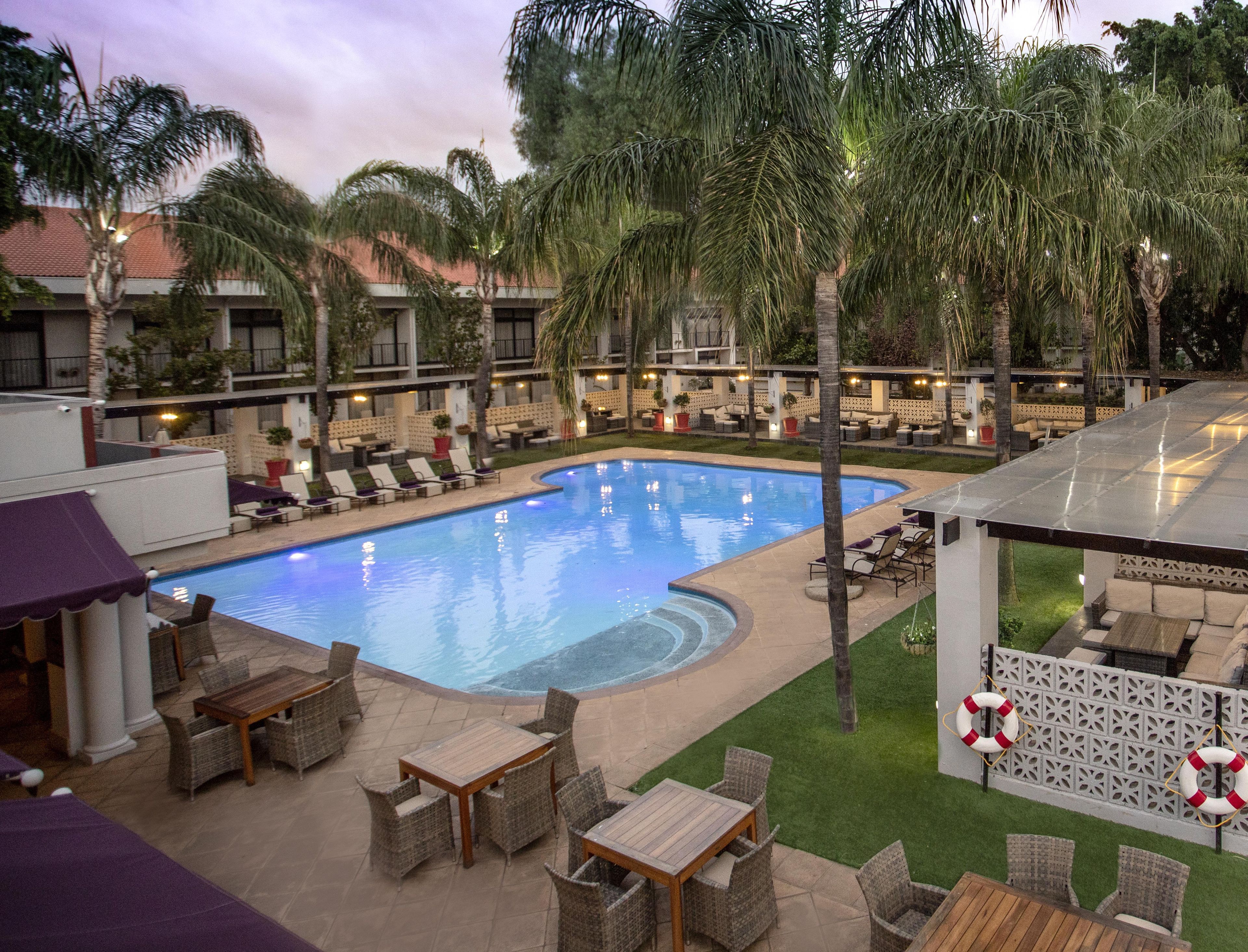 A swimming pool surrounded by lounge chairs and tables, with palm trees and a building in the background.