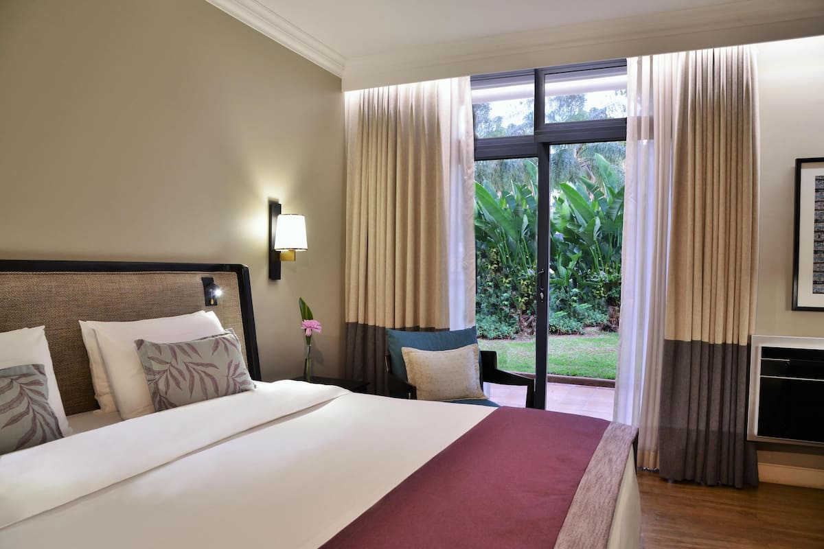 Une chambre d’hôtel avec un grand lit, une table de chevet avec une lampe, une chaise et une vue sur la verdure par la fenêtre.