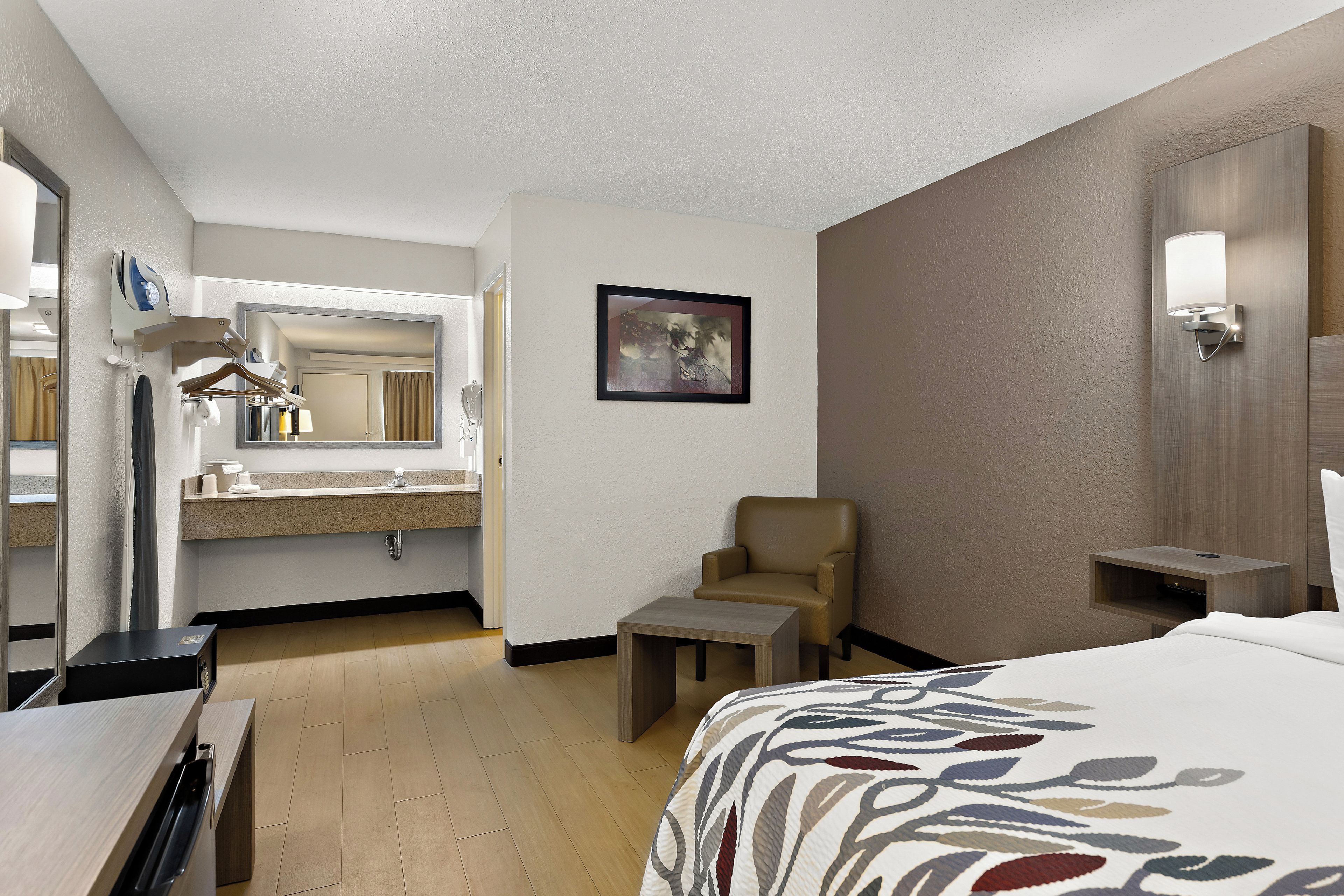 A hotel room with a bed, a chair, a desk, a mirror, a painting, and a bathroom with a sink and mirror.