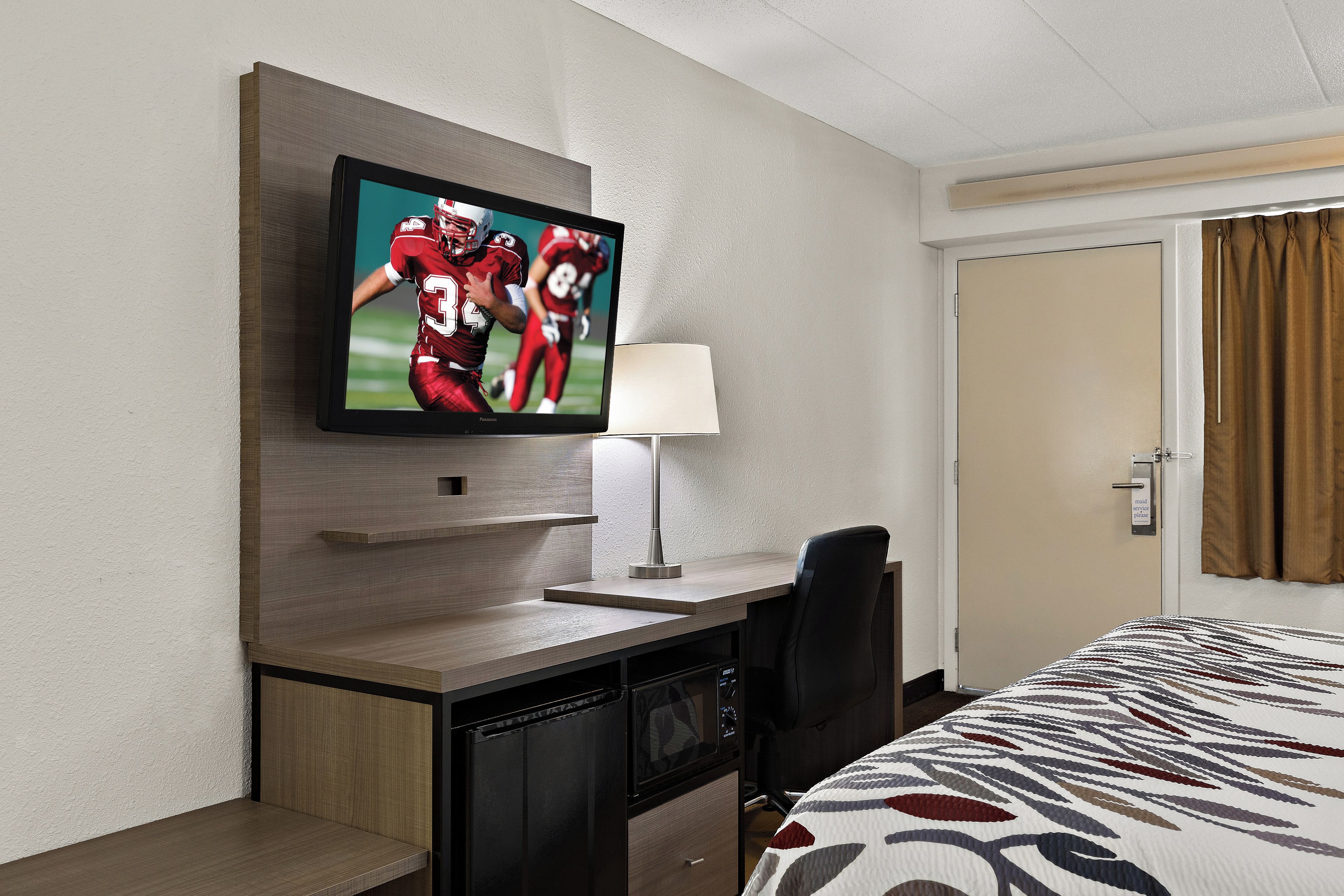 A hotel room with a flat-screen TV mounted on the wall, a desk with a lamp, a chair, and a bed with a patterned bedspread.