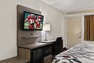 Habitación de hotel con televisor de pantalla plana montado en la pared, un escritorio con lámpara, una silla y una cama con cubrecama estampado.