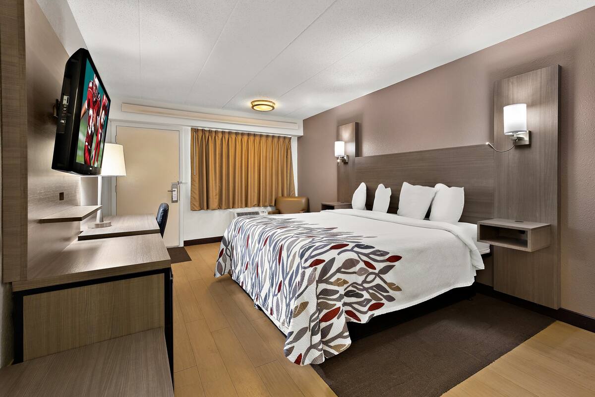 A modern hotel room with a large bed, a desk, a flat-screen TV, and a wall-mounted lamp.