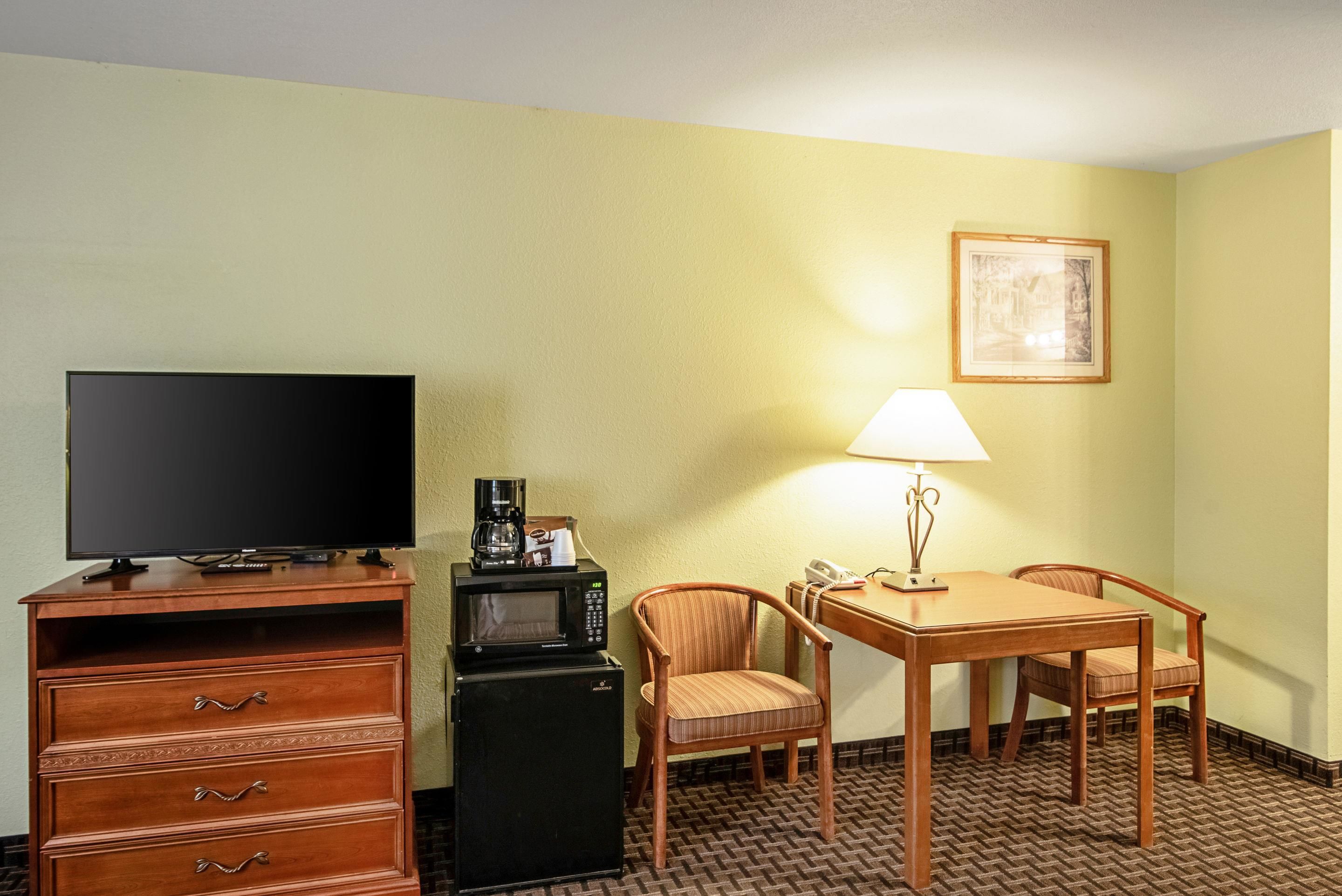 Ein Hotelzimmer mit Flachbildfernseher, einer Kaffeemaschine, einer Mikrowelle, einem Tisch mit zwei Stühlen, einer Lampe und einem gerahmten Bild an der Wand.