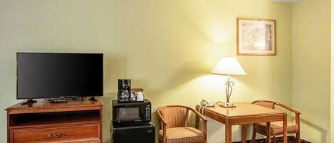 Habitación de hotel con televisión de pantalla plana, cafetera, microondas, mesa con dos sillas, lámpara e imagen enmarcada en la pared.