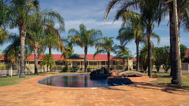 A swimming pool with a brick pathway, palm trees, and a building with a red roof in the background. - Pines Country Club Motor Inn (Shepparton)