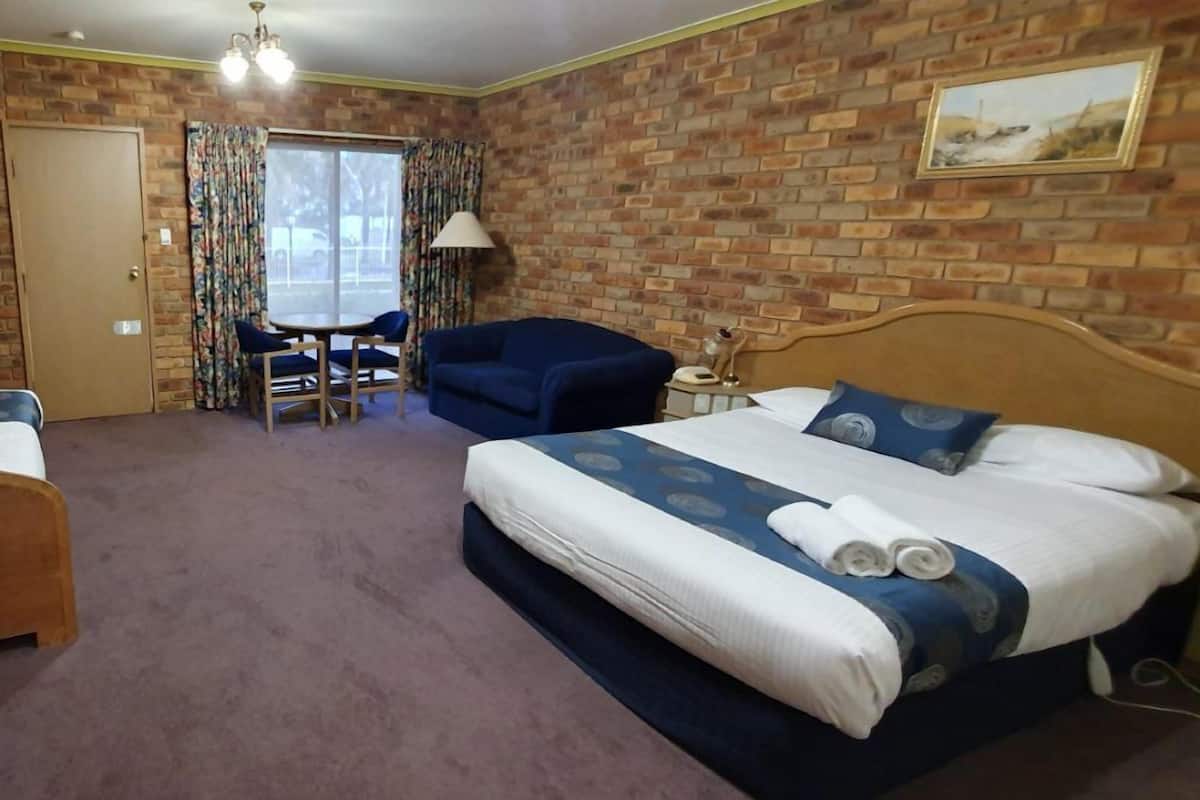 A hotel room with a brick wall, a bed with blue bedding, a blue armchair, a wooden chair, a small table with a lamp, and a door.