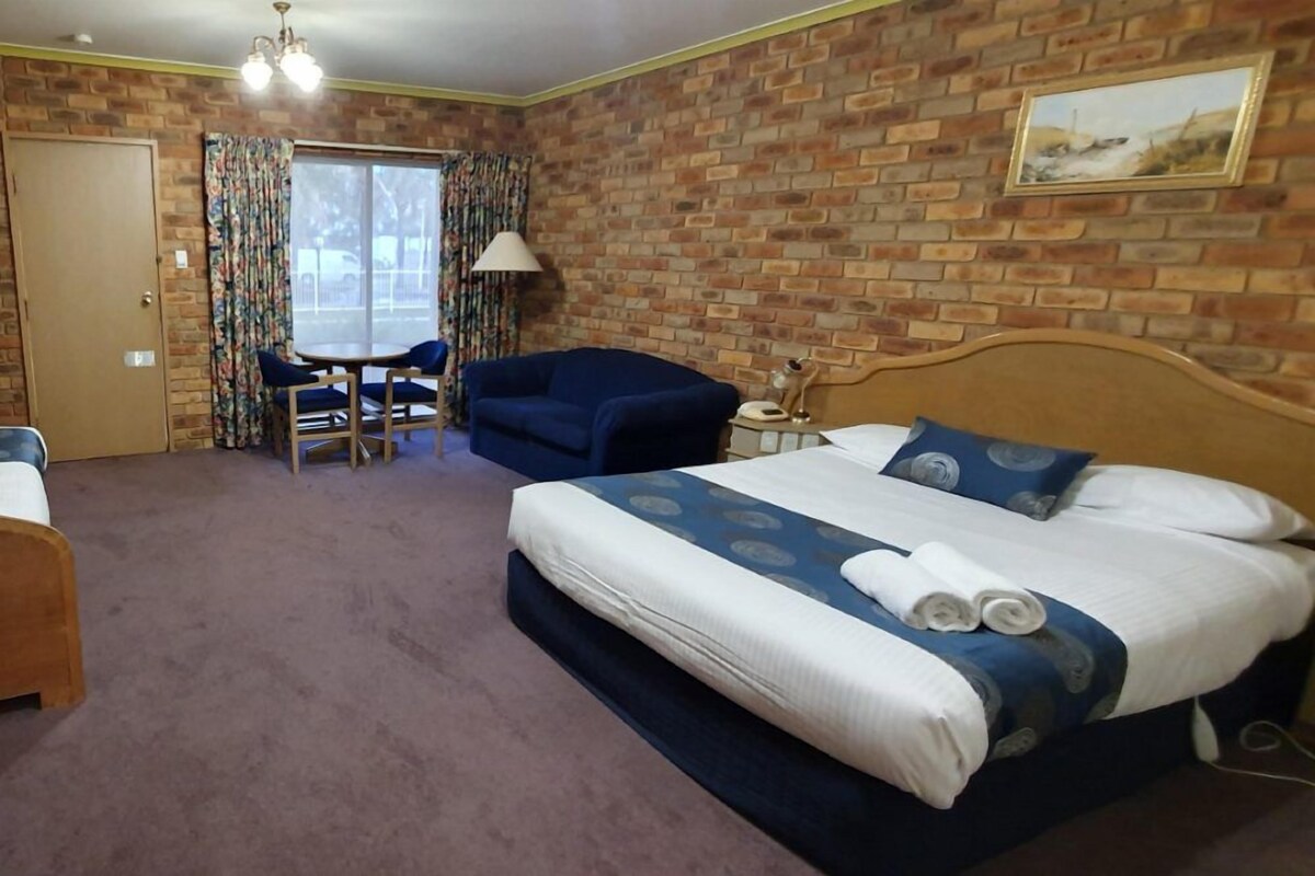 A hotel room with a brick wall, a bed with blue bedding, a blue armchair, a wooden chair, a small table with a lamp, and a door.