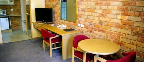 A room with a brick wall, a wooden desk, a television, a mirror, and two red chairs.