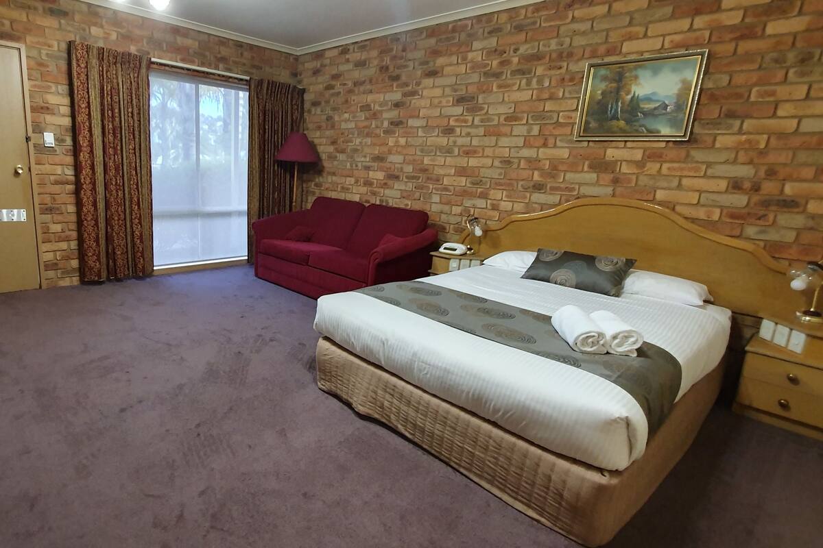 A hotel room with a large bed, a red armchair, a small table, and a sliding door leading to a balcony.