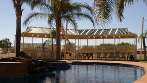 A swimming pool with a covered seating area, palm trees, and a building with a metal roof in the background.