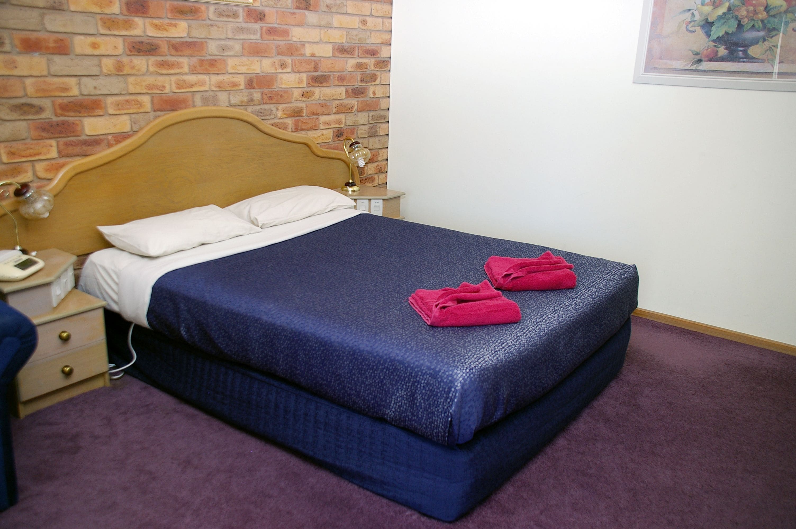 Een net opgemaakt bed met een blauwe sprei en twee rode handdoeken, een houten hoofdbord, een nachtkastje met een lamp en een bakstenen muur.