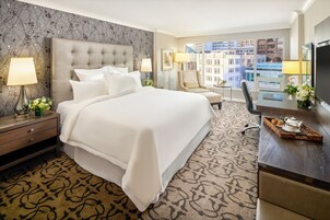 Quarto de hotel com uma cama grande, uma mesa com televisão, uma cadeira e vista para a cidade.