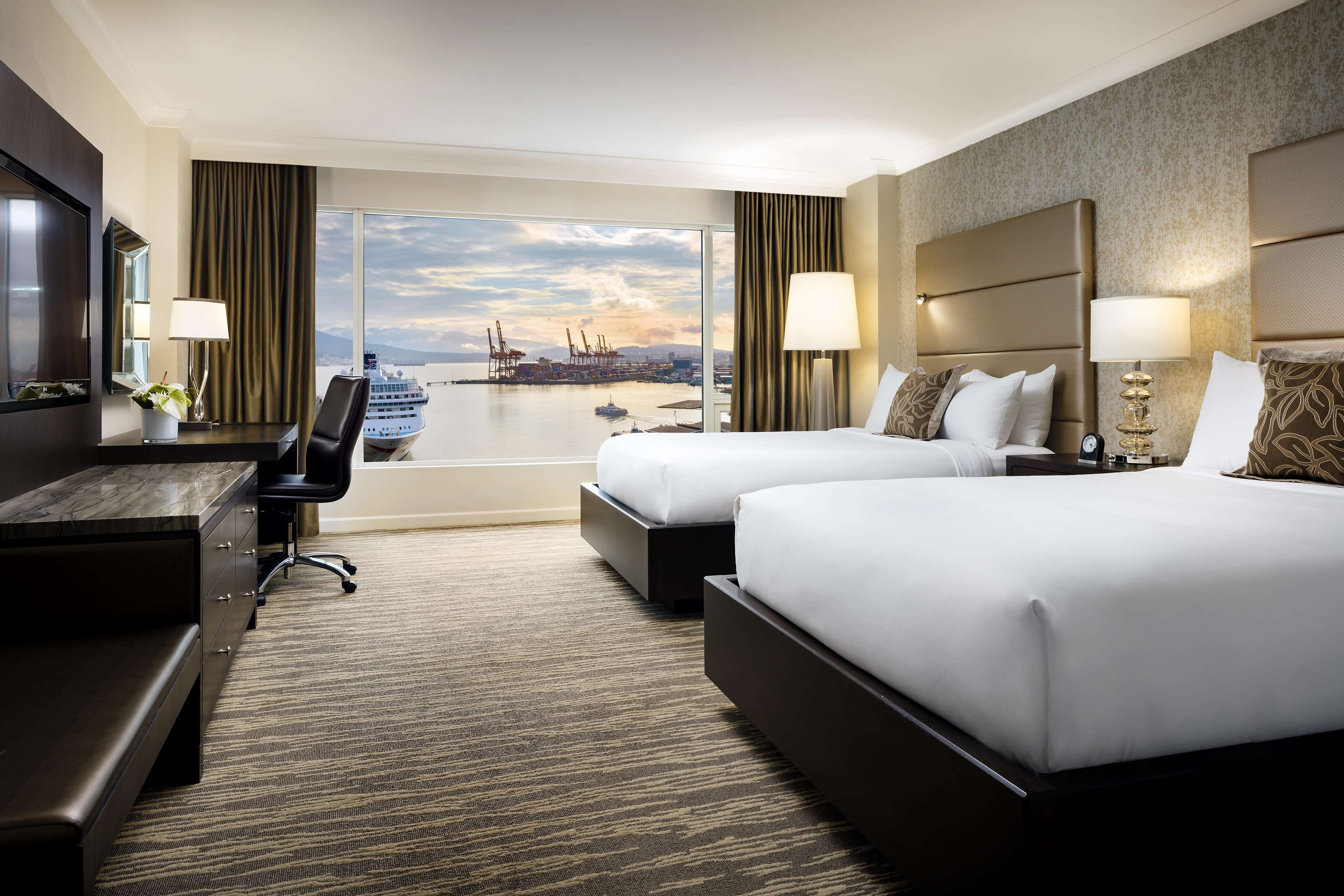 A hotel room with two beds, a desk, and a large window offering a view of a port and ships.