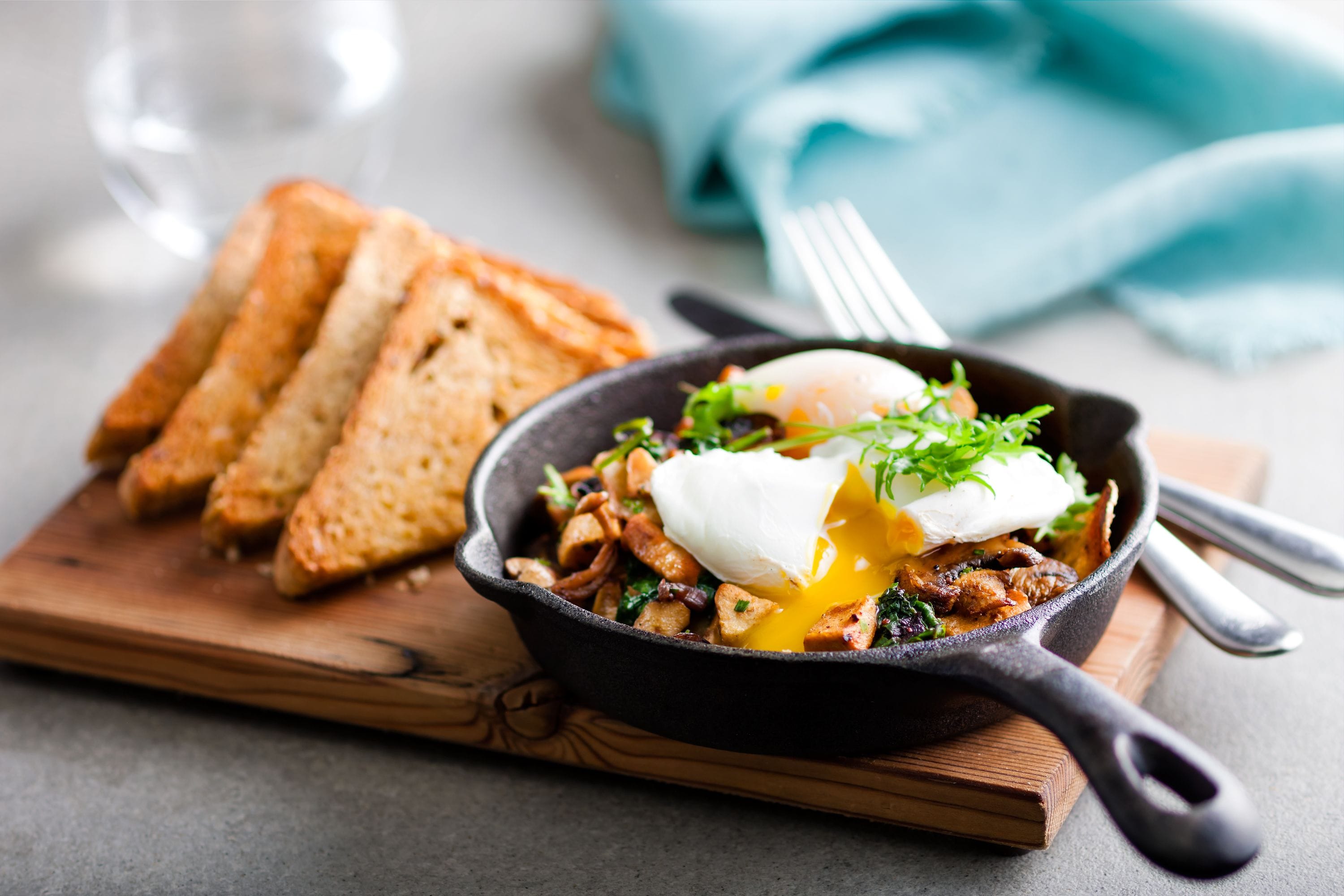 A skillet with a poached egg and vegetables on a wooden board, accompanied by toasted bread slices.