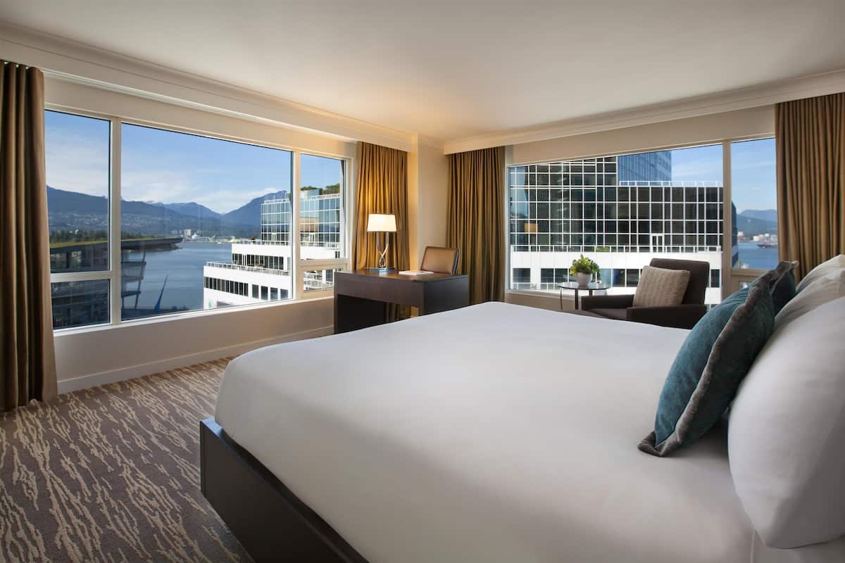 A hotel room with a large bed, a desk with a lamp, a chair, and a view of a river and mountains.
