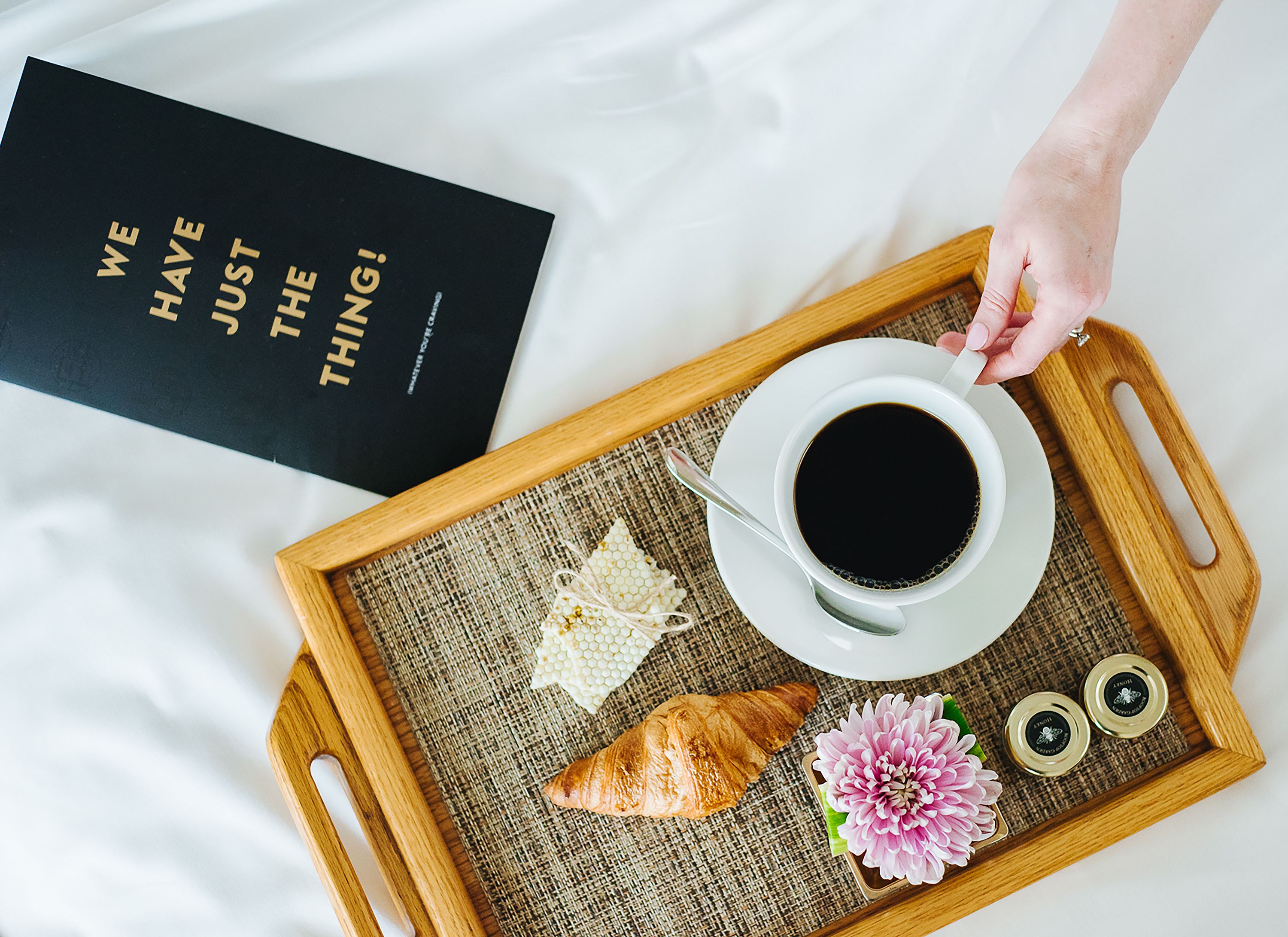 Una bandeja con un croissant, una flor y dos tarros de mermelada, junto con una taza de café y un libro sobre la cama.