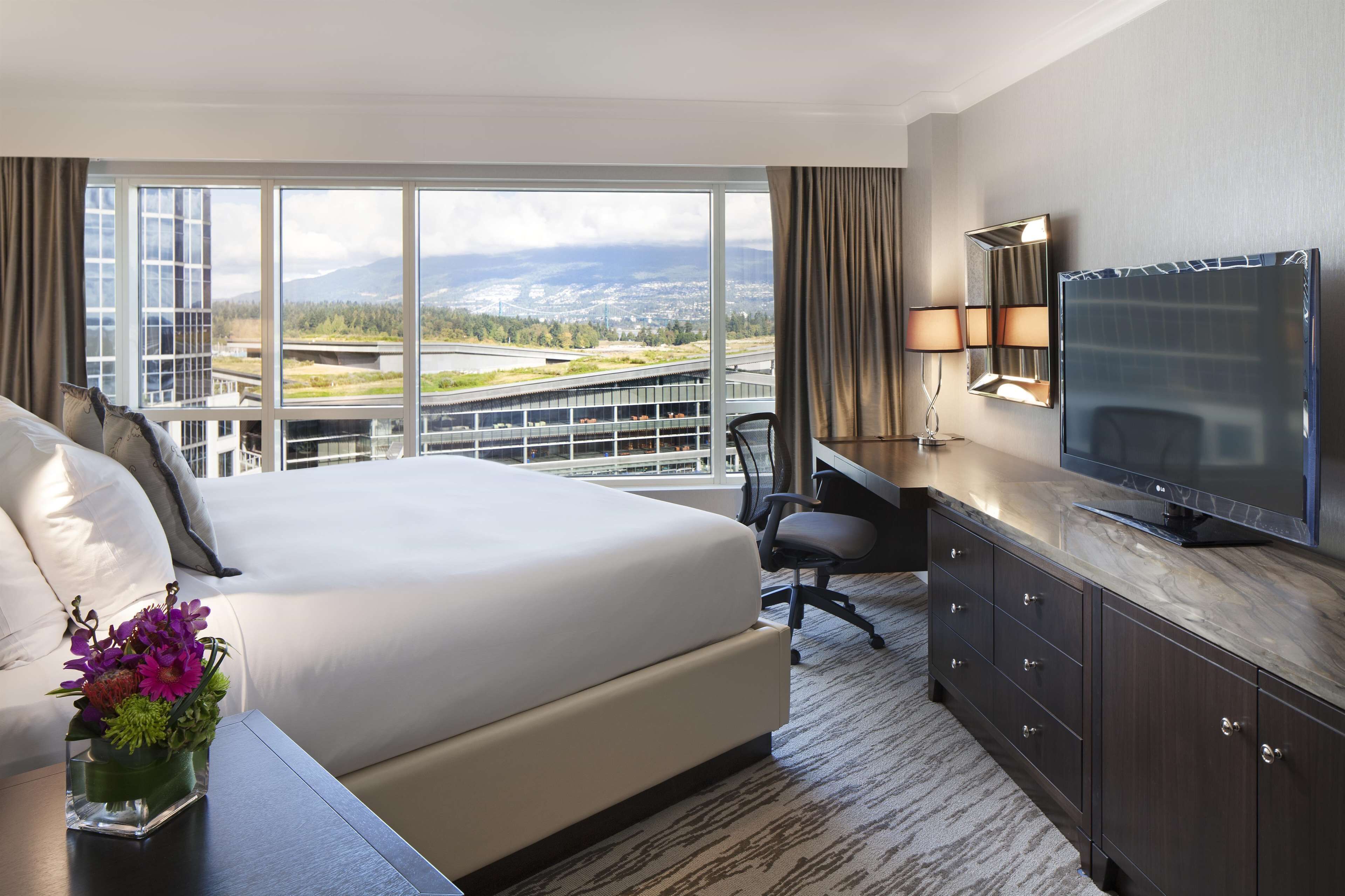 A hotel room with a large bed, a desk with a chair, a television, and a view of a highway and mountains.