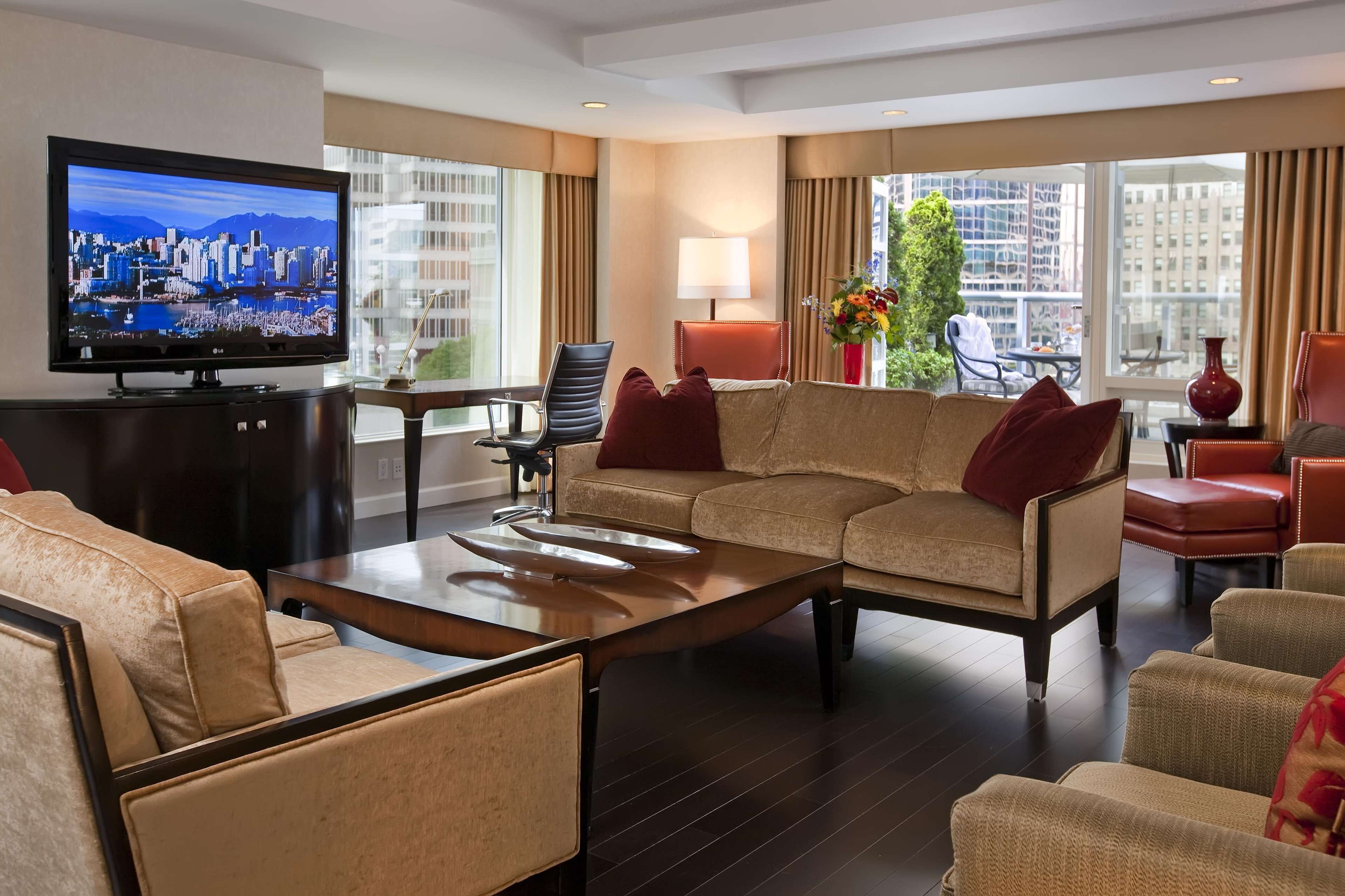 A hotel living room with a large flat-screen TV, a sofa, armchairs, a coffee table, and a view of the city through large windows.