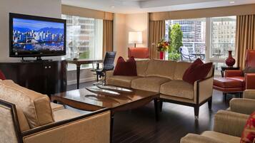 A hotel living room with a large flat-screen TV, a sofa, armchairs, a coffee table, and a view of the city through large windows.