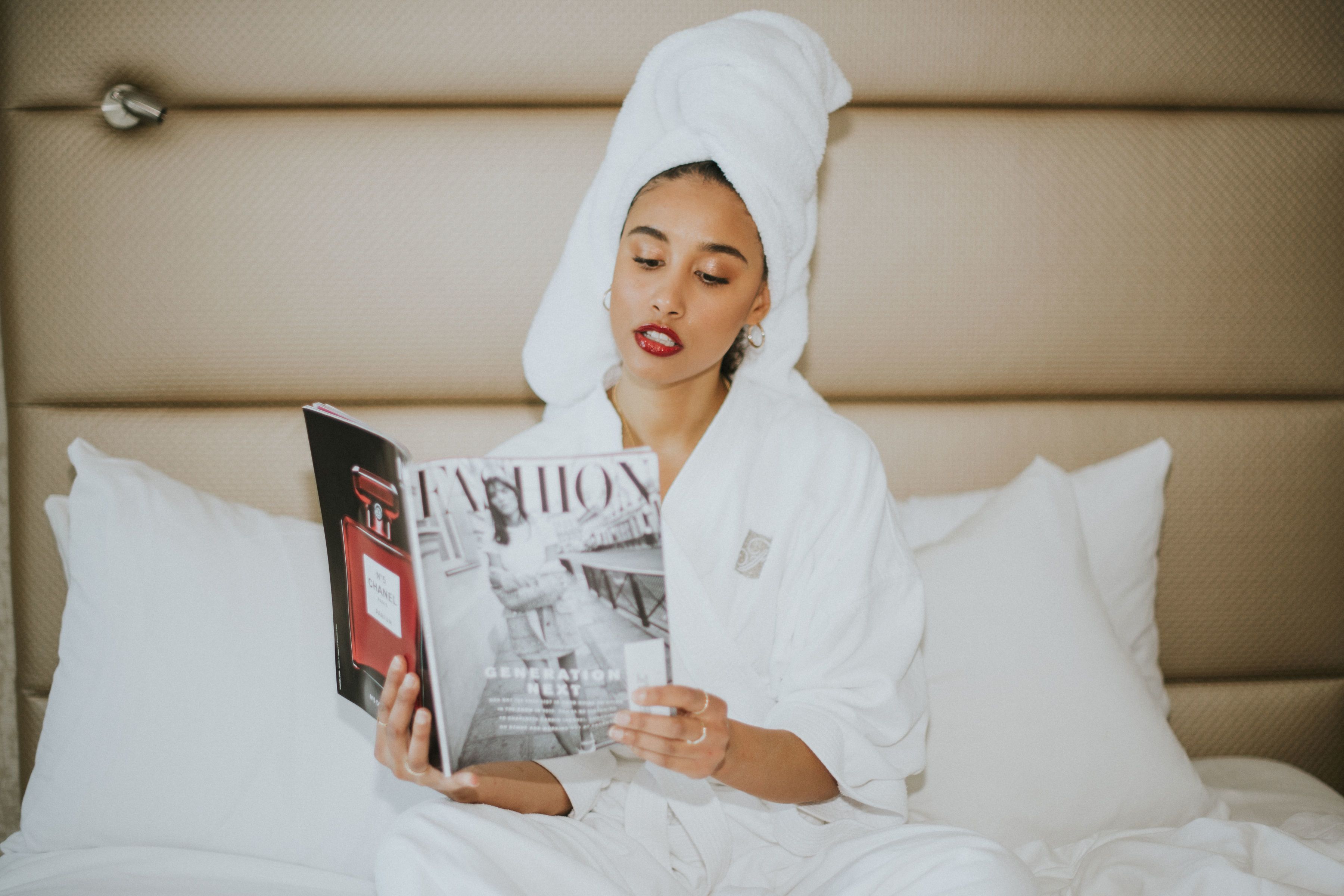 A person in a white bathrobe sitting on a bed with white pillows, reading a magazine.