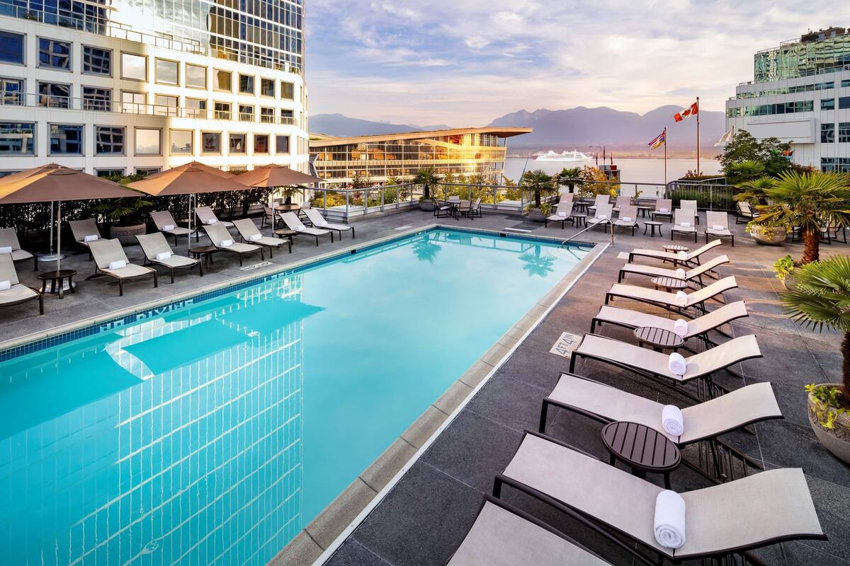 A pool area with lounge chairs and umbrellas, surrounded by buildings with large windows and a view of the sea.