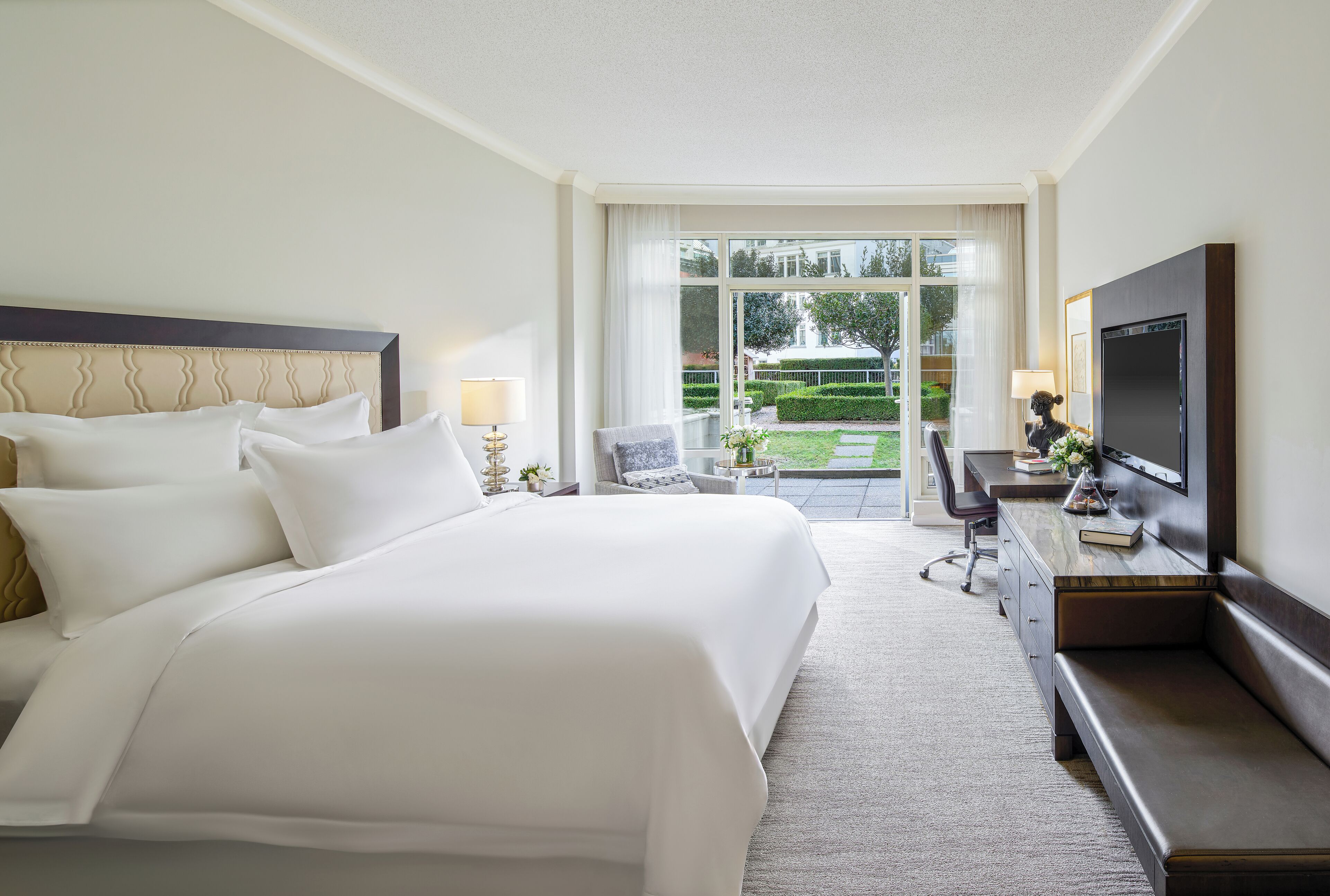 Habitación de hotel con una cama grande, un escritorio con televisor y vistas a un jardín por una puerta de vidrio.