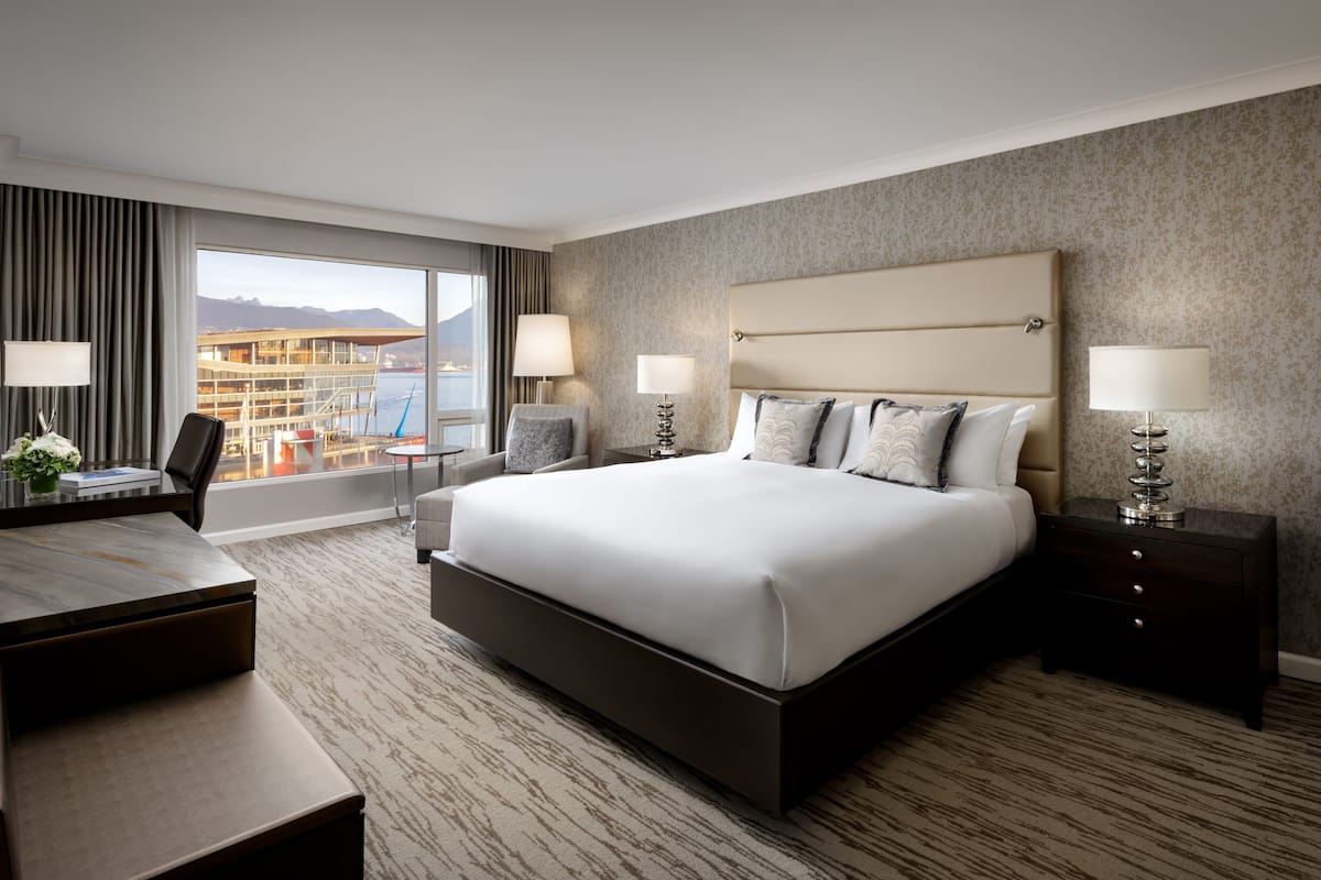 A hotel room with a large bed, two bedside tables with lamps, a desk with a chair, and a view of a building and mountains through the window.