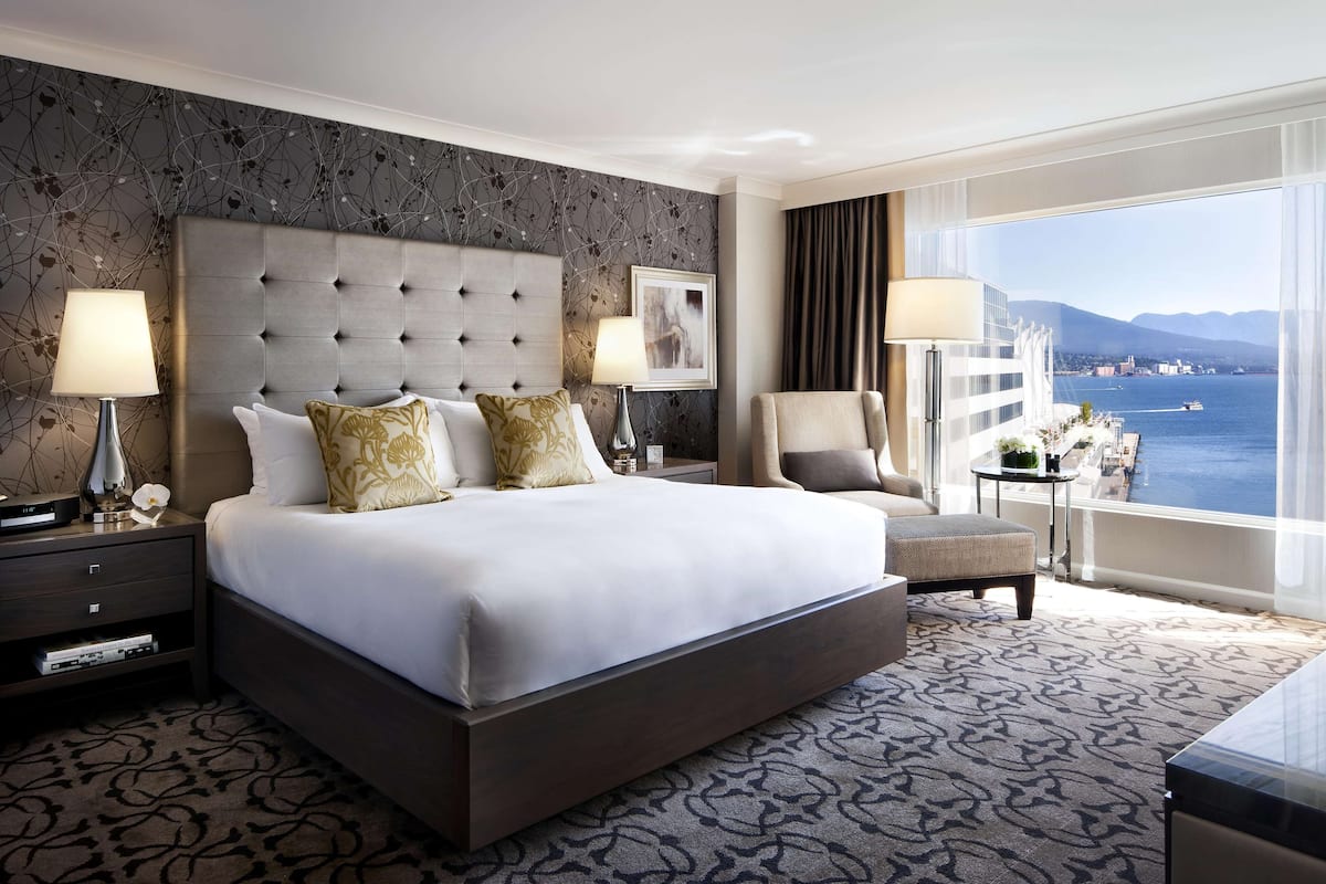 A hotel room with a large bed, two bedside tables, a chair, a view of the sea, and a balcony with a railing.