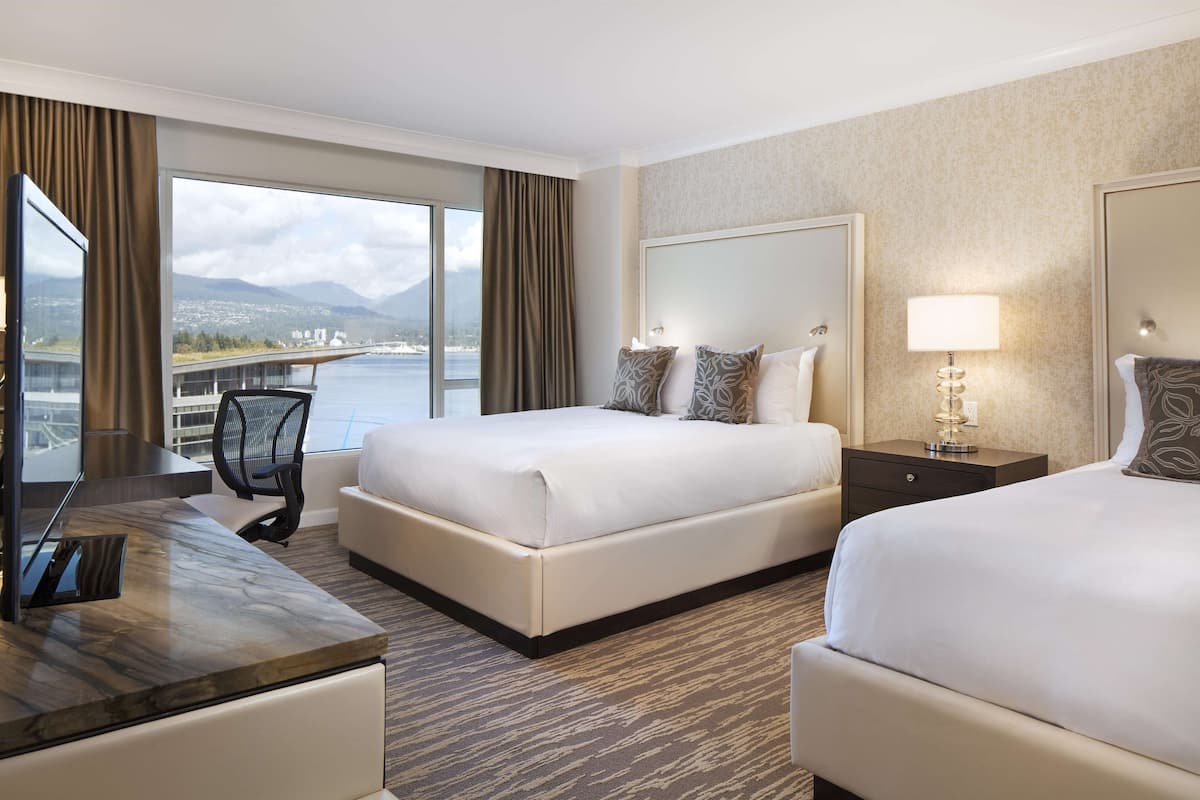 A hotel room with two beds, a desk, and a large window offering a view of a bridge and mountains.