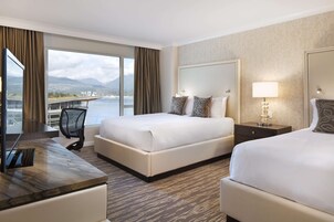 Habitación de hotel con dos camas, un escritorio y un amplio ventanal que ofrece vistas a un puente y montañas.