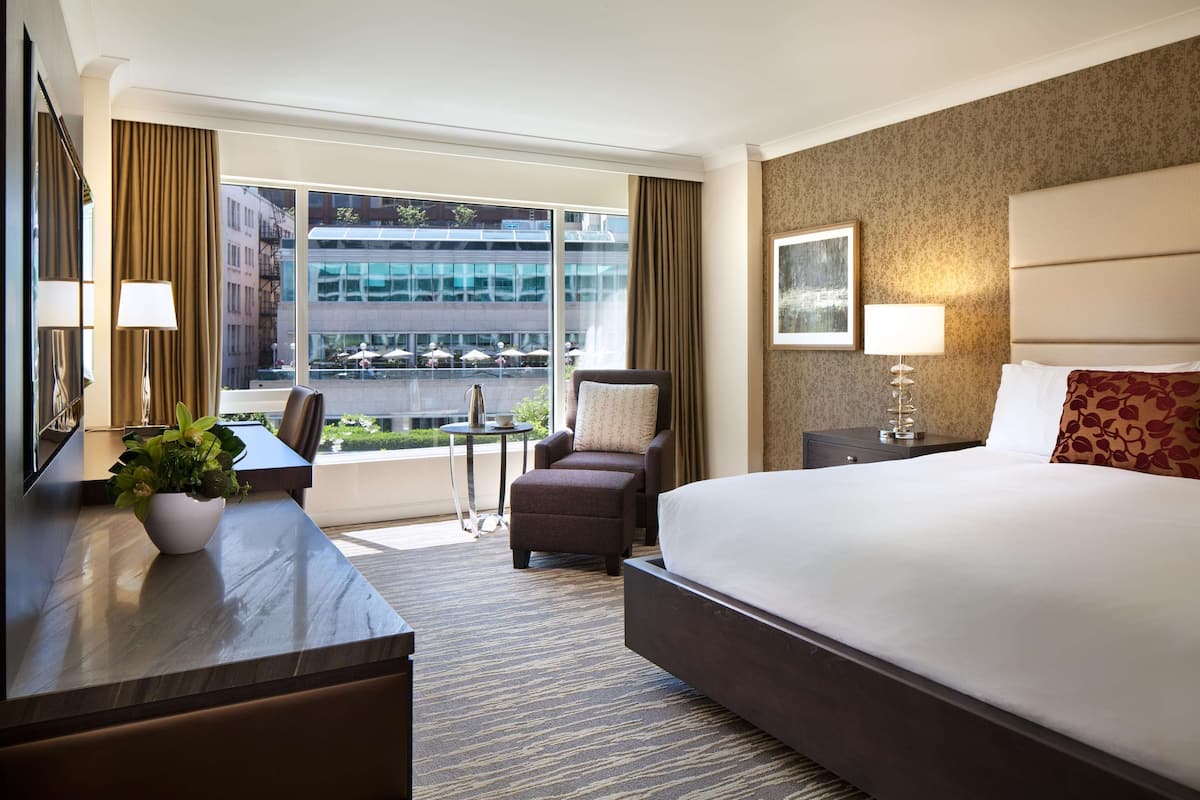 A hotel room with a large bed, a desk with a lamp, a chair, and a view of the city through large windows.