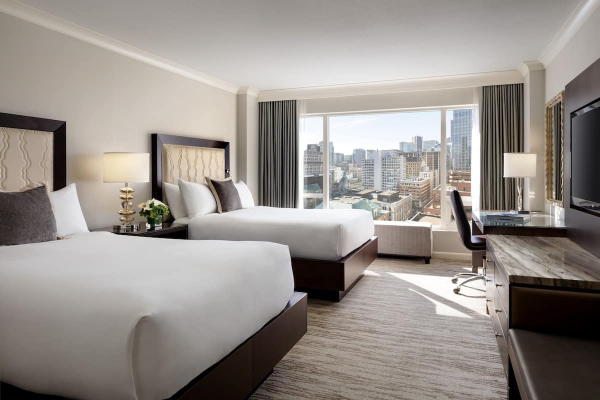 A hotel room with two beds, a desk, a TV, and a large window with a city view.
