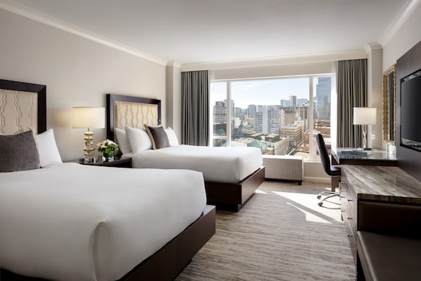 A hotel room with two beds, a desk, a TV, and a large window with a city view.