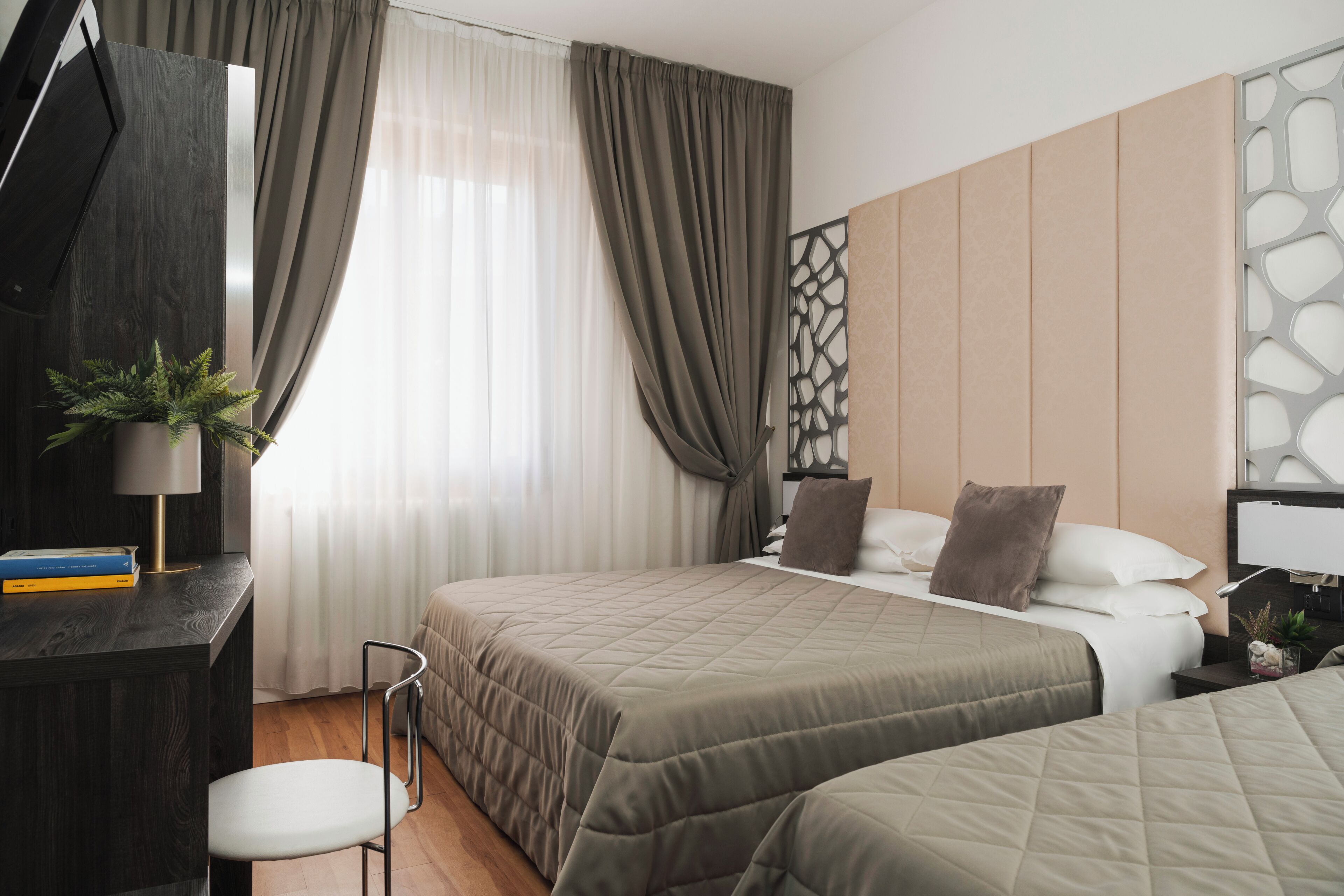 Ein Hotelzimmer mit zwei Betten, einem Schreibtisch, einem Stuhl, einer Zimmerpflanze und einem Vorhangfenster.
