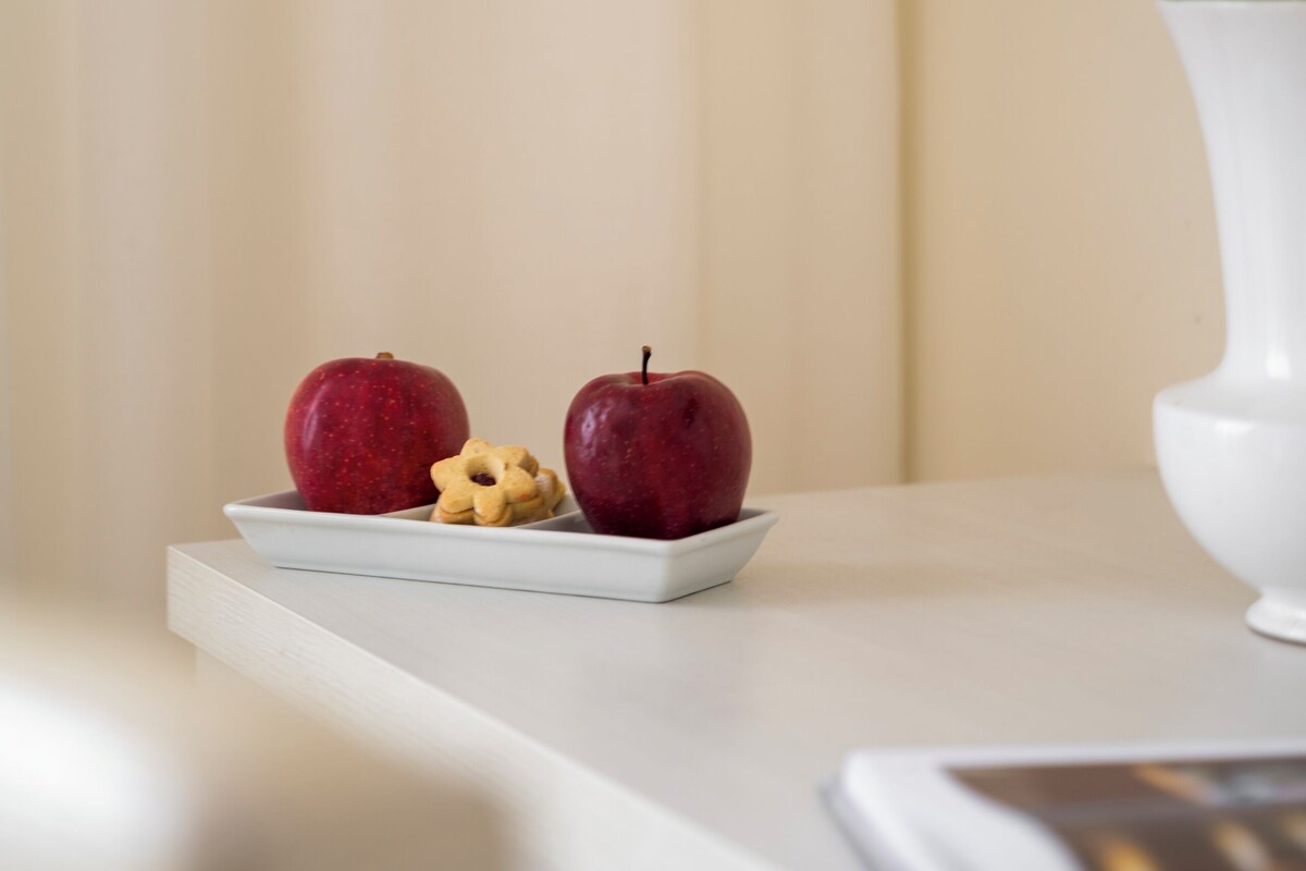 Un jarrón blanco con una orquídea blanca sobre una mesa blanca, junto a un plato blanco con dos manzanas rojas y una galleta.