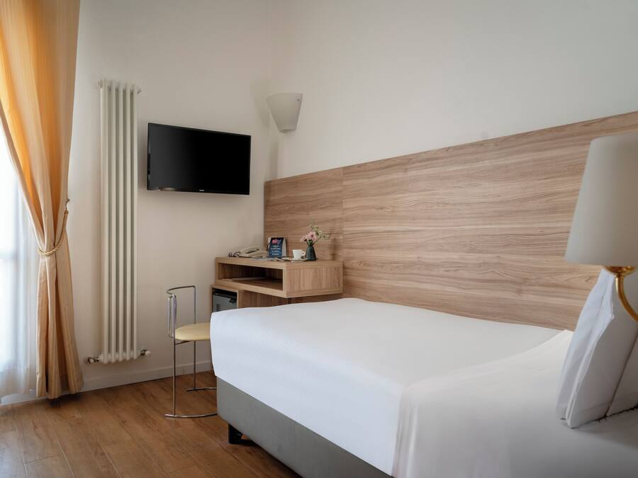 Una camera d'albergo con un letto, una televisione a parete, un comodino con lampada e una finestra con tende.