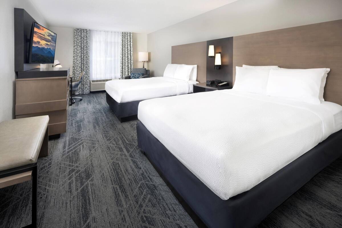 Quarto de hotel com duas camas, uma escrivaninha, uma cadeira, uma televisão e uma janela com cortinas.