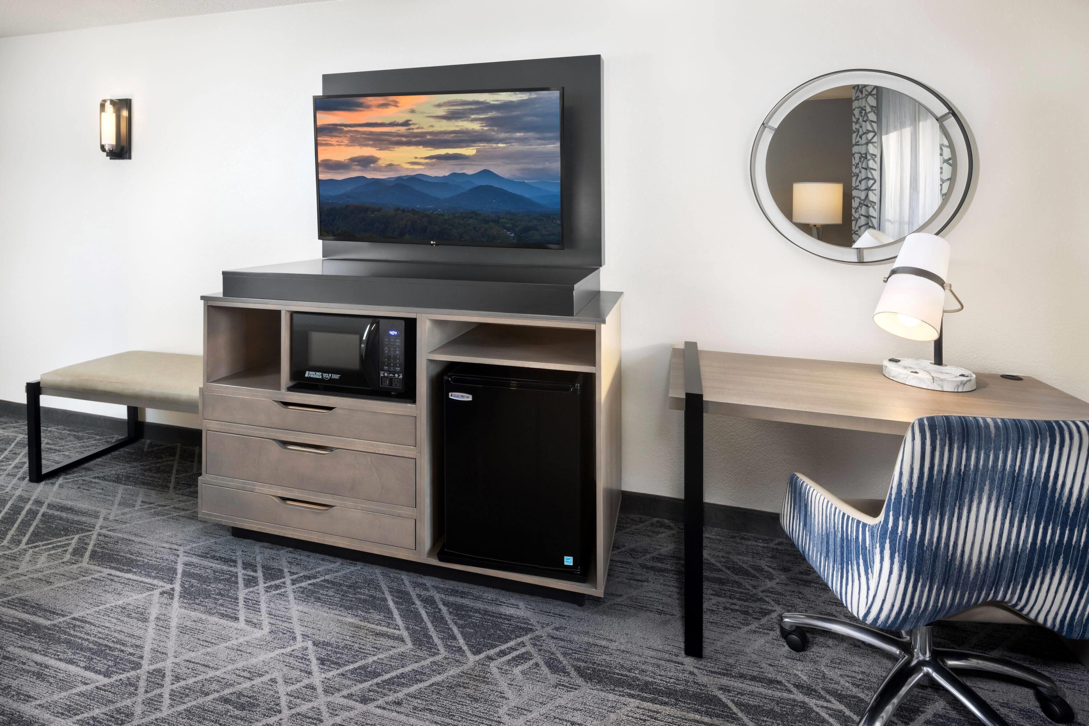 Habitación de hotel con televisor de pantalla plana en la pared, un escritorio con lámpara, una silla y un minibar.