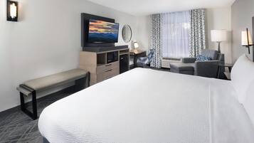 A hotel room with a large bed, a desk with a chair, a bench, a television, and a window with curtains.