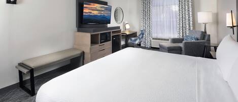 Habitación de hotel con una cama grande, un escritorio con silla, un banco, un televisor y una ventana con cortinas.