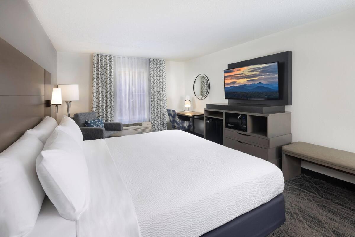 Quarto de hotel com uma cama grande, uma mesa com cadeira, televisão e janela com cortinas.