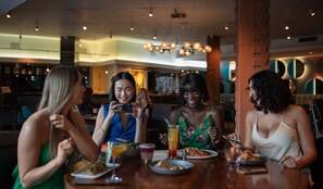 A group of people dining at a restaurant with a modern interior, including a brick column and pendant lighting. - Hotel Valley Ho (Scottsdale)