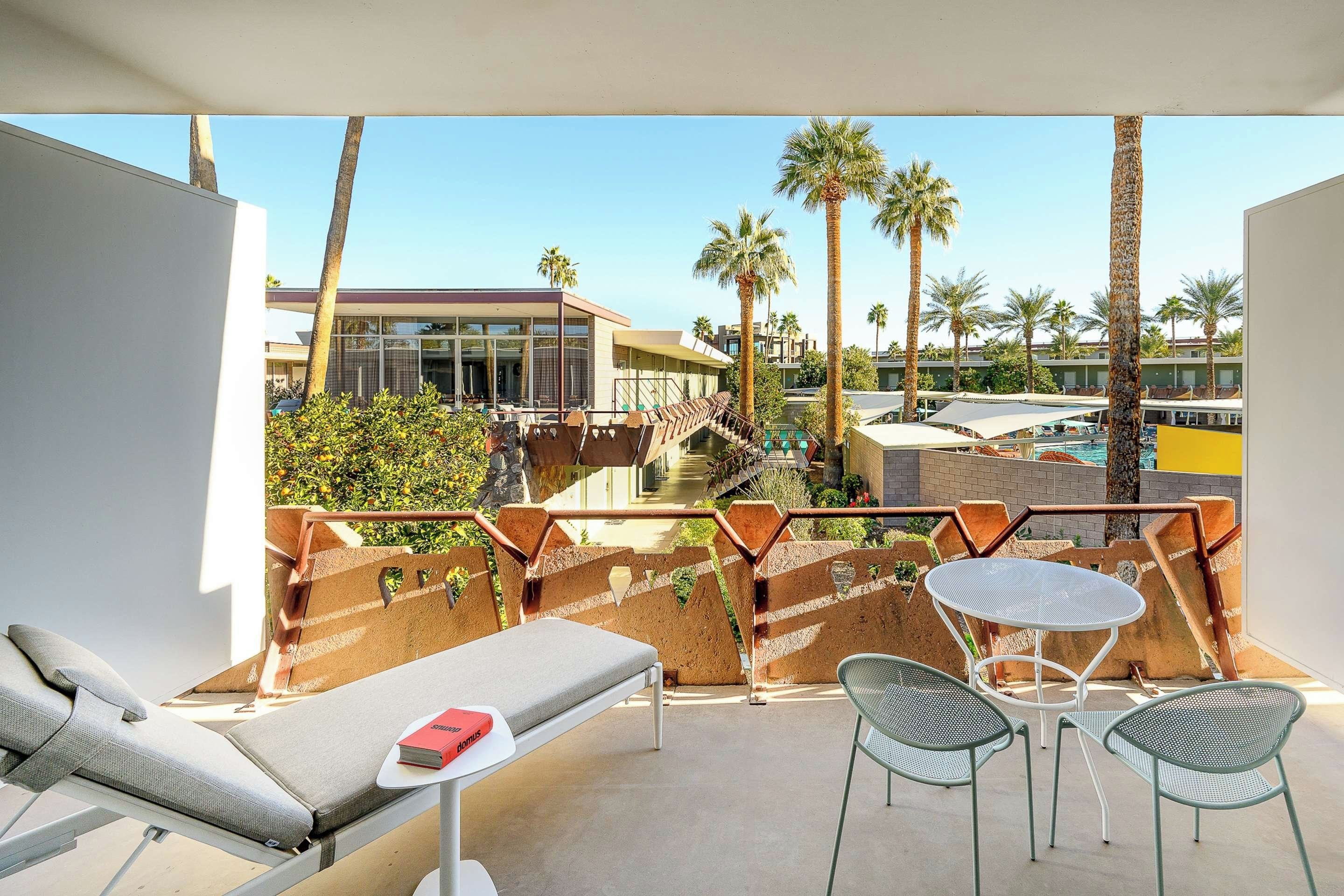 A balcony with a lounge chair, a small table, and a chair. There are palm trees and a building in the background.
