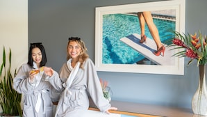 Two individuals in bathrobes holding drinks, a framed picture of a poolside scene, and a potted plant on a reception desk. - Hotel Valley Ho (Scottsdale)