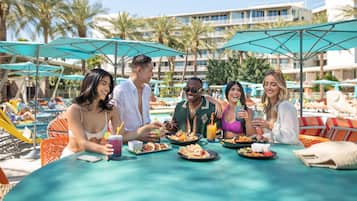 A group of people dining at an outdoor table with umbrellas, surrounded by palm trees and a pool.