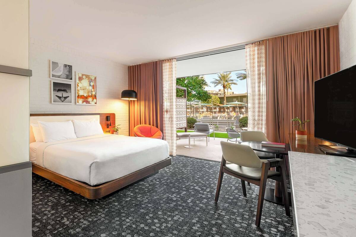 A hotel room with a bed, a desk, a chair, a TV, and a view of the outdoor area with a pool and palm trees.