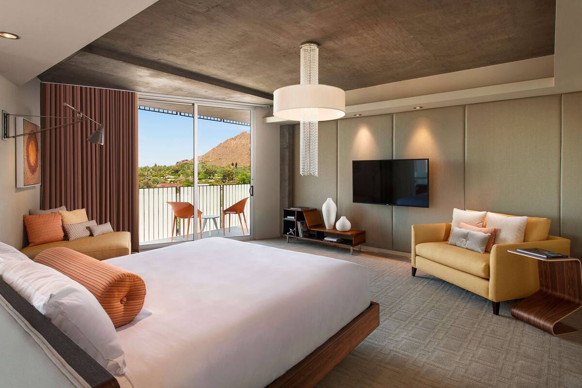 A modern hotel room with a large bed, a sofa, a TV, and a balcony with a mountain view.