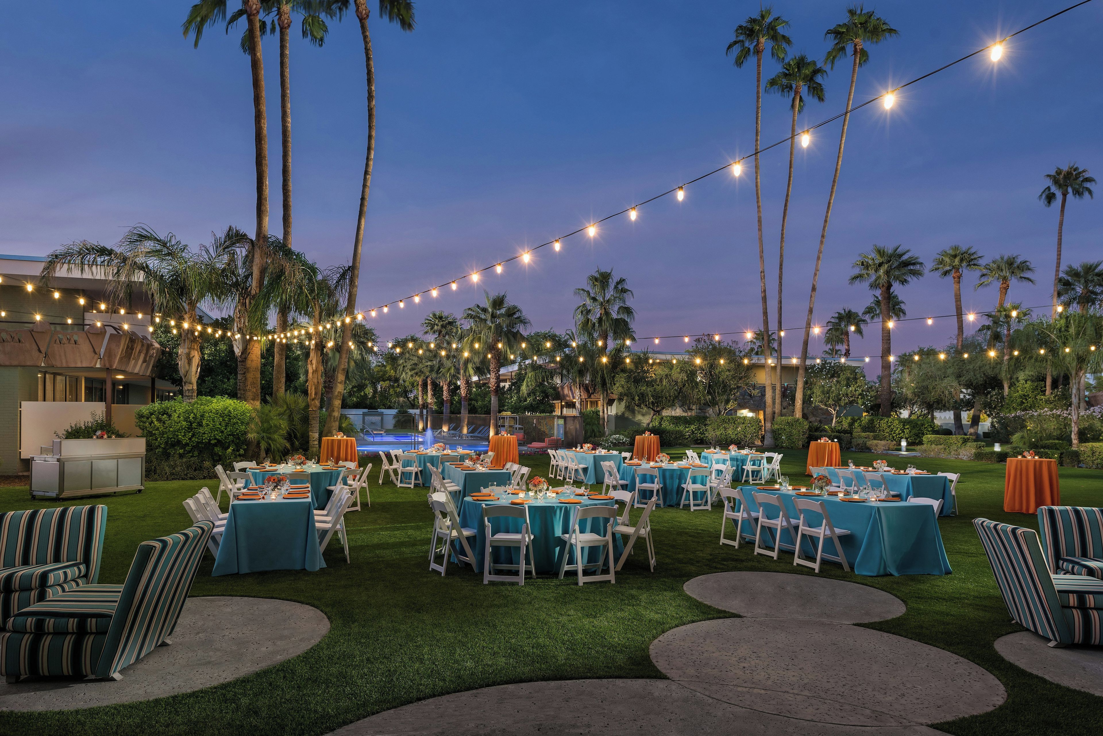 An outdoor event space with tables set for dining, surrounded by palm trees and string lights.