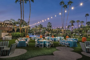 An outdoor event space with tables set for dining, surrounded by palm trees and string lights.